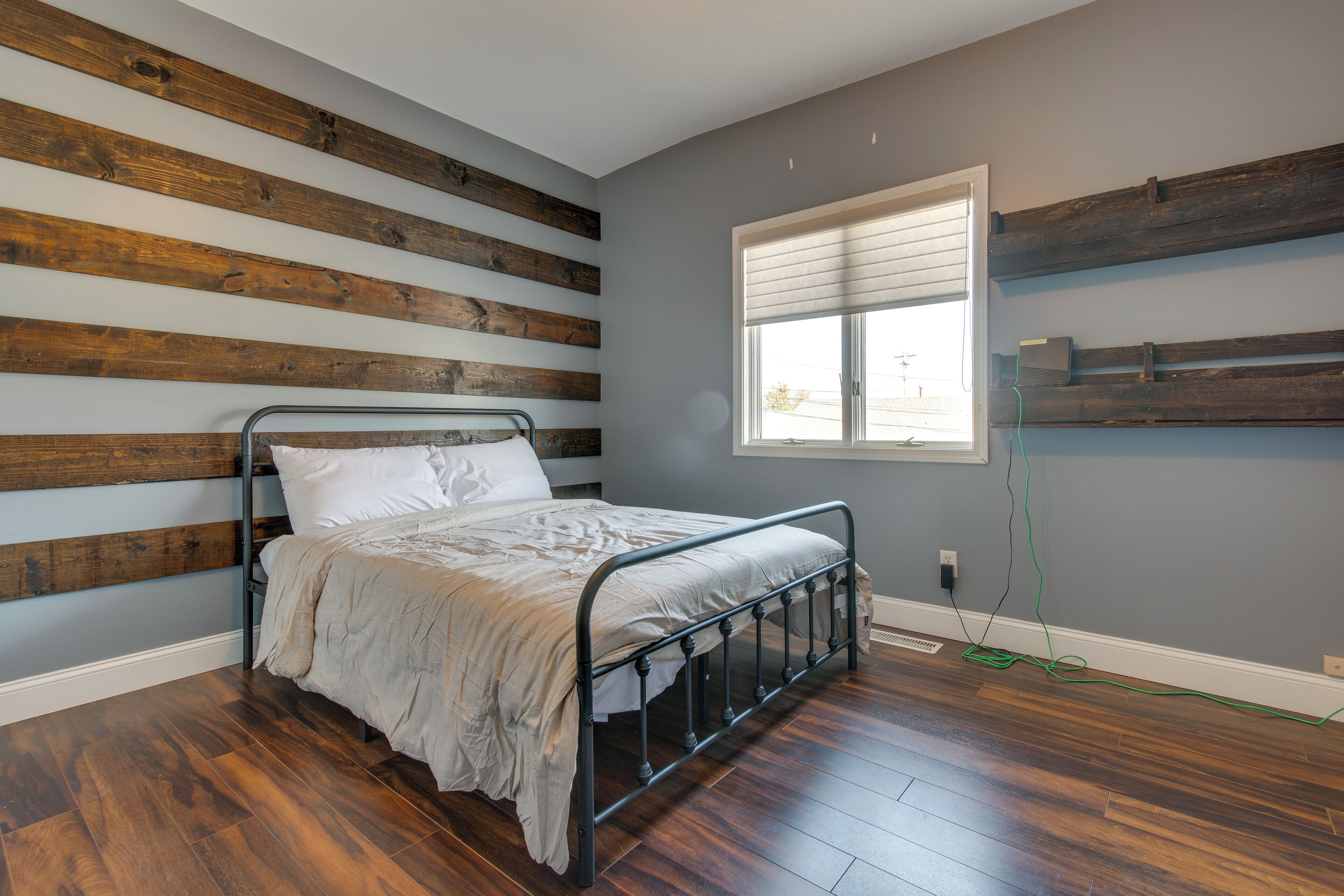 Quarto com estrutura de cama de metal, ripas de madeira na parede, janela com persianas e uma tomada.