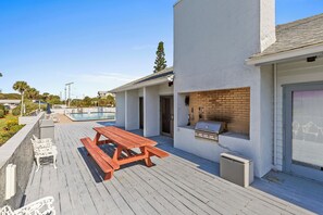 Une terrasse en bois avec des tables de pique-nique, un barbecue et une piscine en arrière-plan.