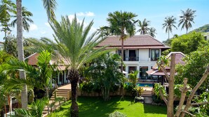 A two-story house with a swimming pool, surrounded by lush greenery and palm trees.