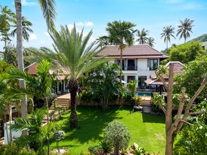 Uma casa de dois andares com piscina, rodeada por vegetação exuberante e palmeiras.