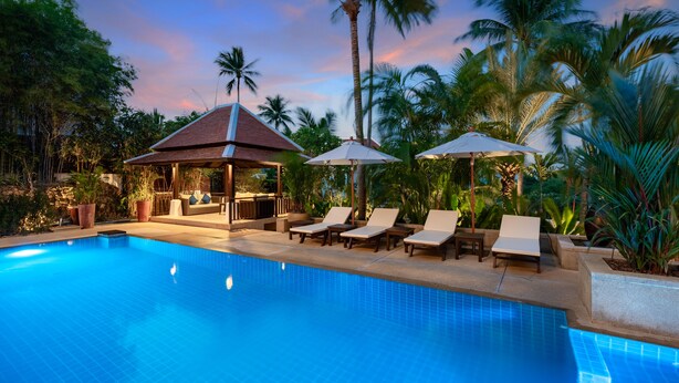 A swimming pool with lounge chairs and umbrellas, surrounded by palm trees and a gazebo.