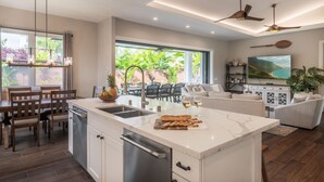 A modern kitchen with a central island, dining area with wooden chairs, and a living room with a sofa and TV. - Hale Ho'okipa: 2 Homes/2 Pools in Poipu-Sleeps 16 (Koloa)