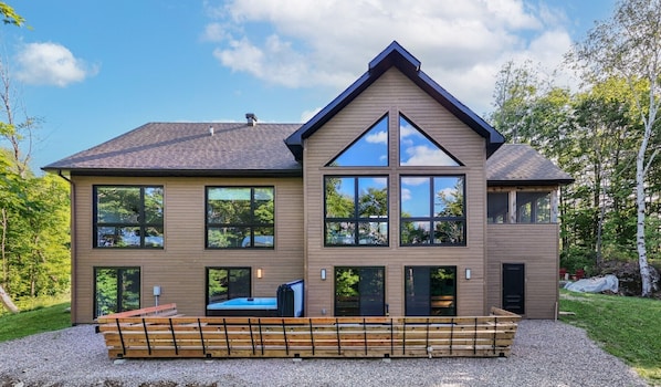 Une maison moderne à deux étages, dotée de grandes fenêtres, d’un patio couvert et entourée d’un cadre naturel.