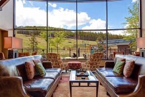 A spacious living area with leather sofas and patterned armchairs, large windows offering a view of a green landscape and a building with solar panels. - Crystal Peak Expedition Condo: Ski-In, Ski-Out Luxury (Breckenridge)