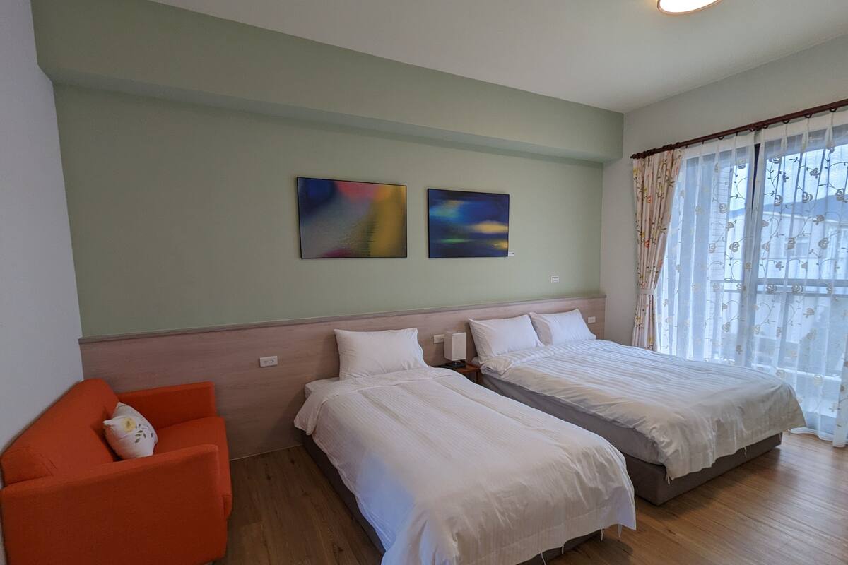 A hotel room with two beds, a small orange chair, and two abstract paintings on the wall.