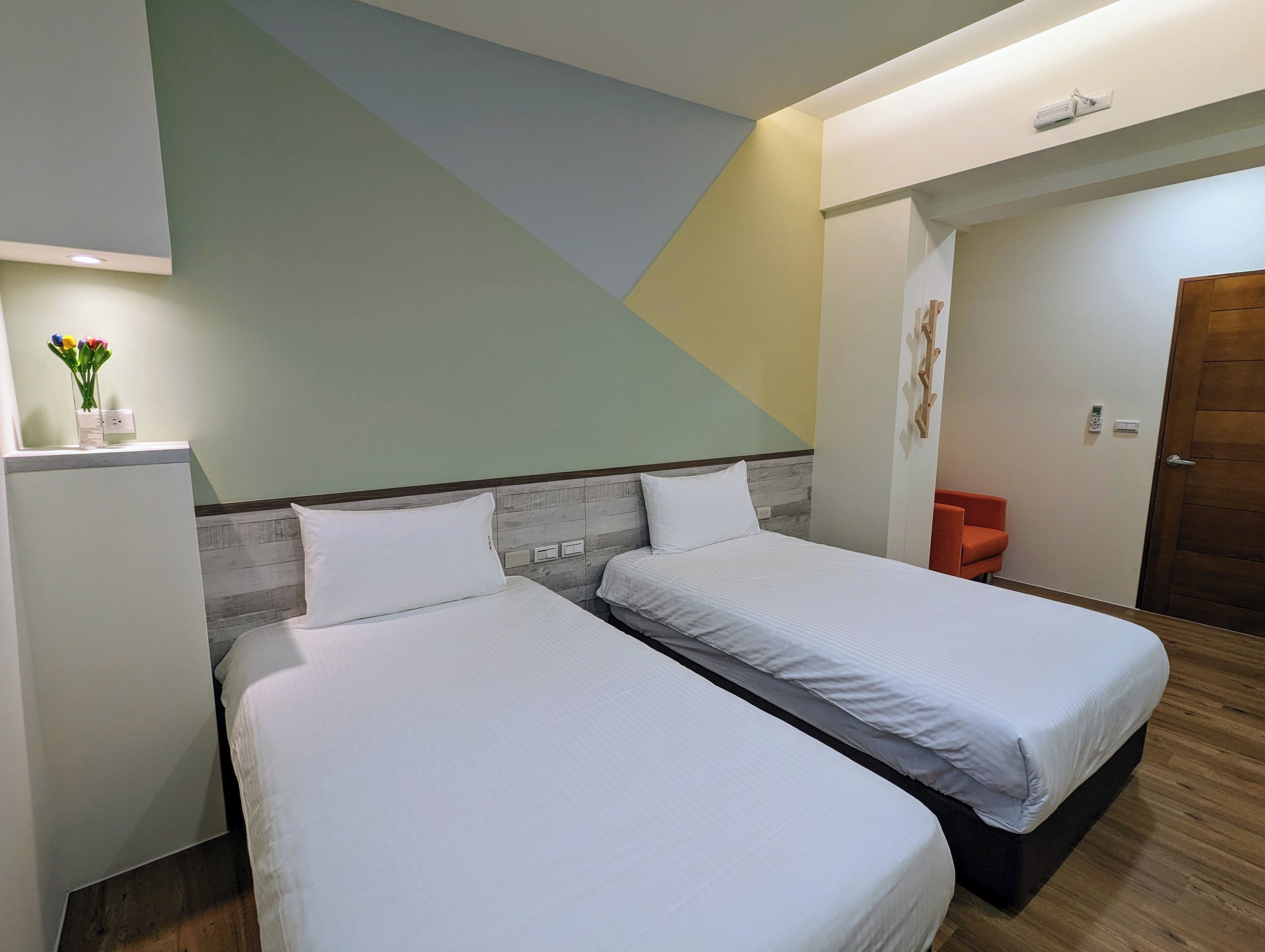 A hotel room with two single beds, a wooden headboard, a wall-mounted air conditioner, a small table with a vase of flowers, and a door with a wooden frame.