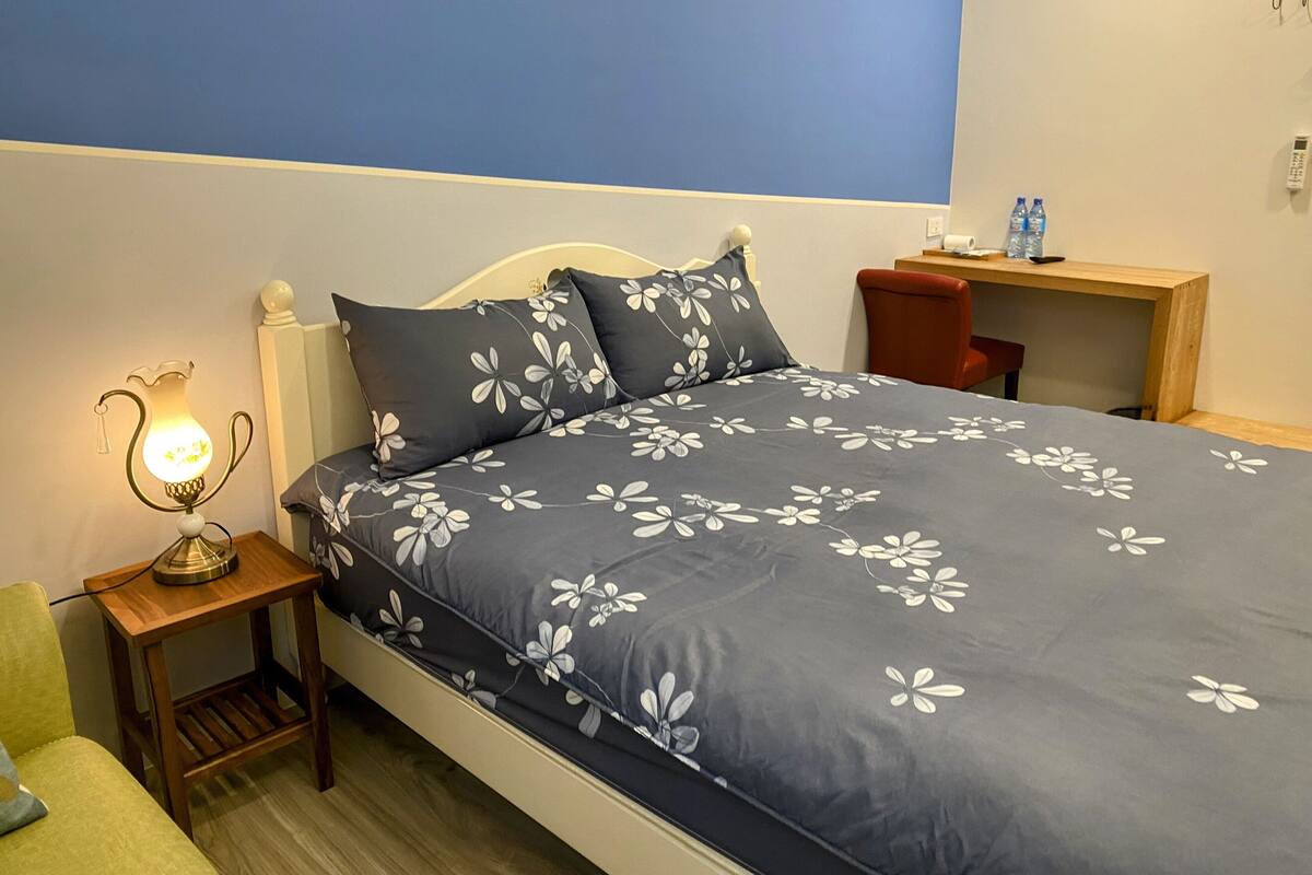 A neatly made bed with a floral duvet, a bedside table with a lamp, and a small wooden nightstand.