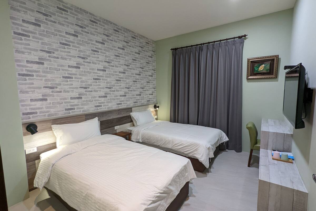 A room with two beds, a brick-patterned wall, a grey curtain, a framed picture, and a TV.