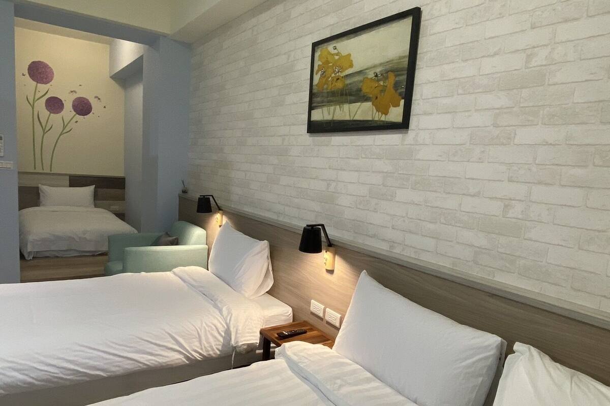 A hotel room with two beds, a light-colored brick wall, and a painting of flowers.
