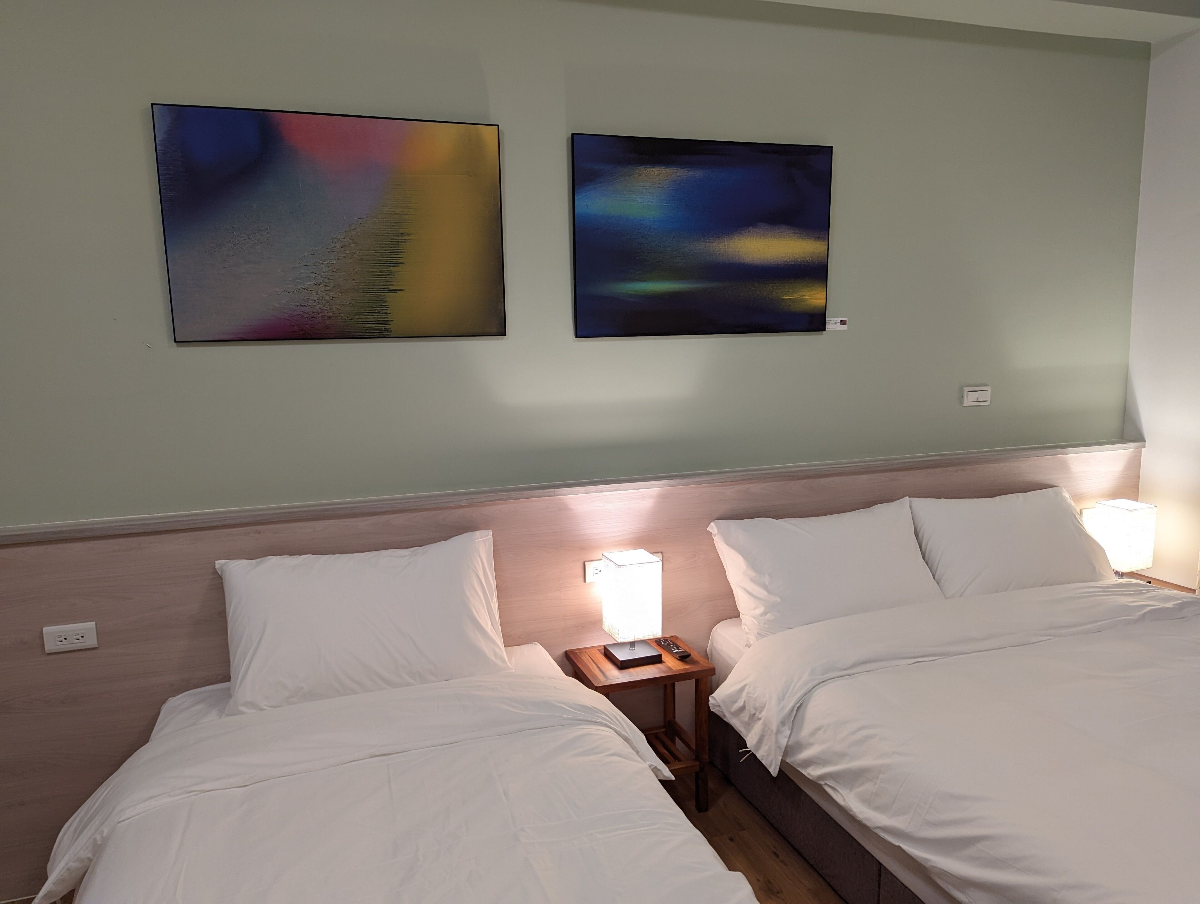 Een hotelkamer met twee bedden, een nachtkastje en twee abstracte schilderijen aan de muur.