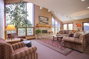 A spacious living room with a stone fireplace, large windows, and a high ceiling.