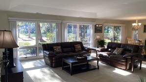 A spacious living room with large windows, a dark brown leather sofa, a coffee table, and a lamp. - Harbour Town Golf Links 11th Tee Ranch Walkable to Harbour Town (Hilton Head Island)