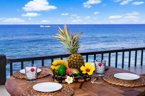 A wooden table set with plates, a pineapple, bananas, a flower, and glasses of water with ice cubes, overlooking the ocean. - Banyan Tree 301: DIRECT OCEANFRONT 2/2 REMODELED WITH AIR CONDITIONING! (Kailua Kona)