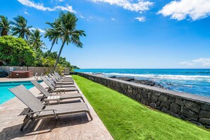 A poolside area with lounge chairs, a stone wall, and a view of the ocean. - Banyan Tree 301: DIRECT OCEANFRONT 2/2 REMODELED WITH AIR CONDITIONING! (Kailua Kona)