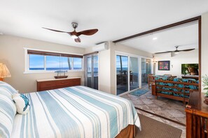 A bedroom with a bed, a ceiling fan, a window with a view, a sliding door to a balcony, and a TV on a wooden stand. - Banyan Tree 301: DIRECT OCEANFRONT 2/2 REMODELED WITH AIR CONDITIONING! (Kailua Kona)