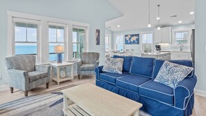 Un salón amplio con un sofá azul grande, dos sillones y una mesa de centro. Hay ventanas grandes que ofrecen vistas al océano.