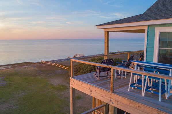 Une terrasse en bois avec des sièges extérieurs, offrant une vue sur une plage et l’océan.