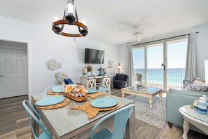 A dining area with a table set for four, a ceiling fan, a television, and a view of the ocean through large windows.