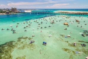 Eine Luftaufnahme einer Küstenlandschaft mit zahlreichen Booten in klarem türkisblauem Wasser, einer in die See führenden Mole und einem weit entfernten Gebäudekomplex.