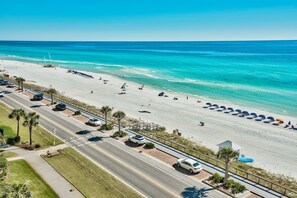 Een luchtendicht van een strand met een weg ernaast, omzoomd door palmbomen en strandparasols.