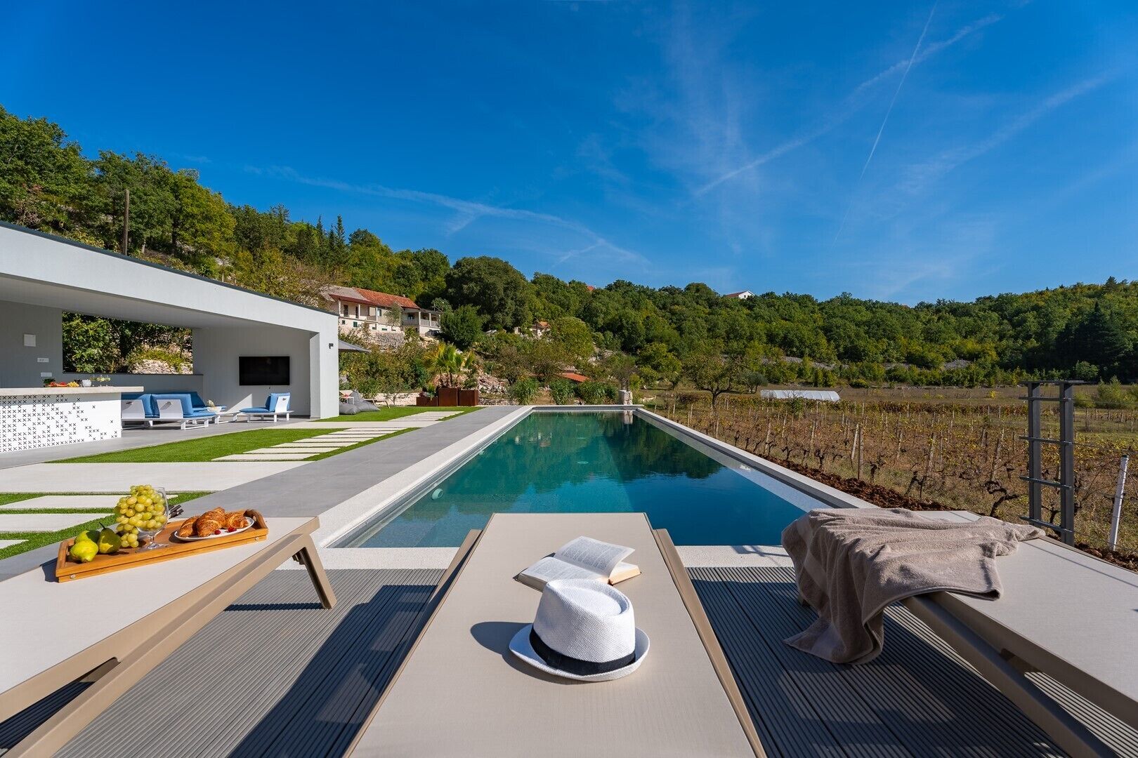 A modern pool area with lounge chairs, a dining table with fruits and pastries, and a white hat on the deck.