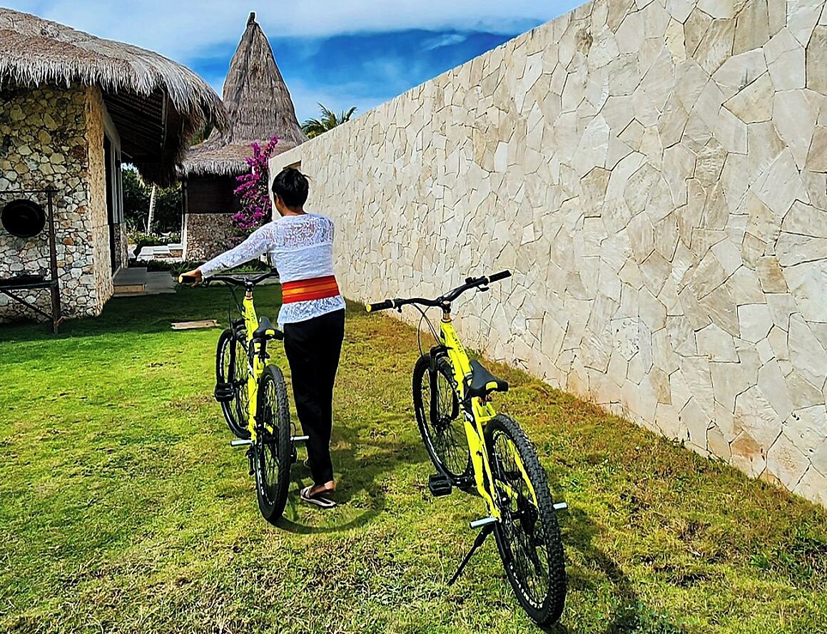 A person standing next to a yellow bicycle in a grassy area with a stone wall and a thatched-roof structure in the background.