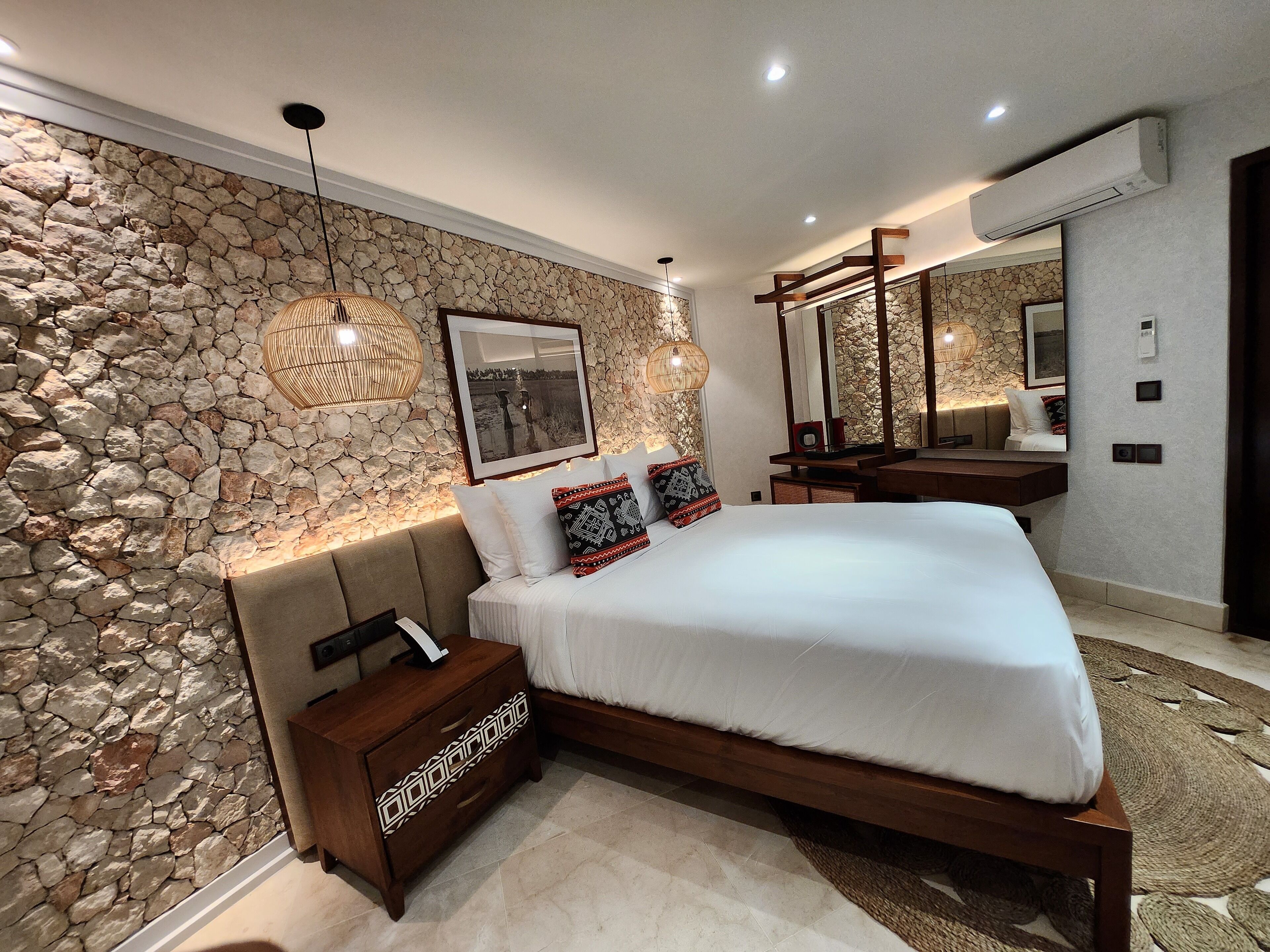 A modern bedroom with a stone wall, a bed with white bedding, a wooden nightstand, and a large mirror.