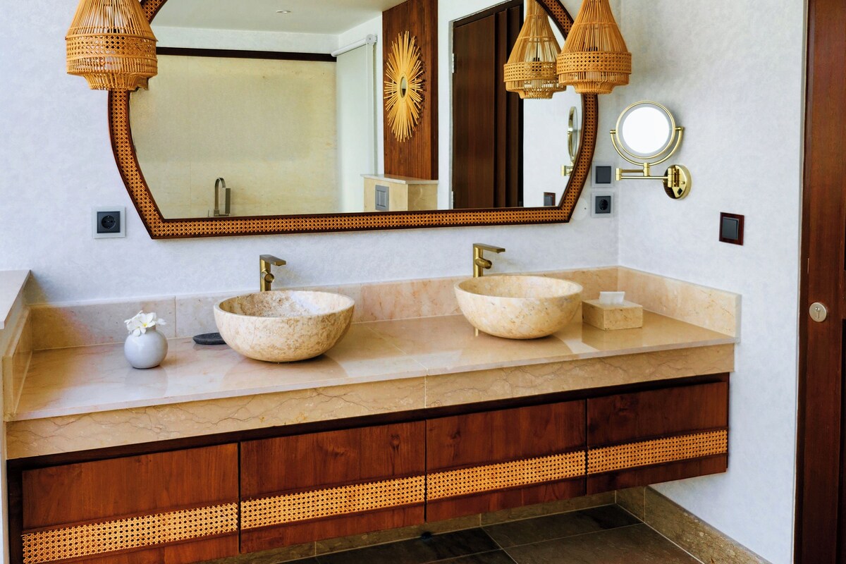 A bathroom with two large, round, beige sinks, a wooden vanity with a woven design, and a large mirror with a decorative frame.