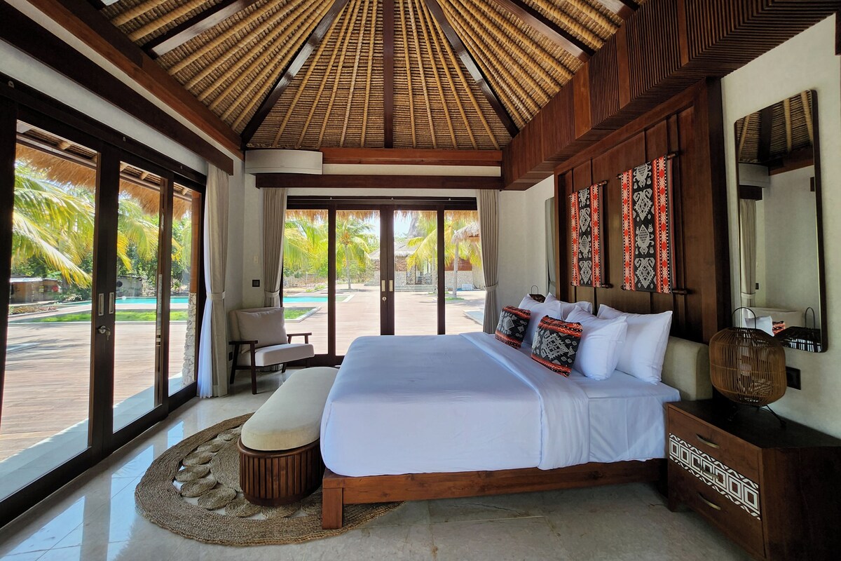A spacious bedroom with a large bed, a wooden ceiling, and a view of a pool and palm trees.