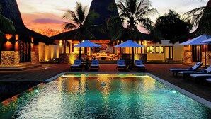 Zona de piscina con sillones de playa y sombrillas, rodeada de palmeras y un edificio con techo de paja.
