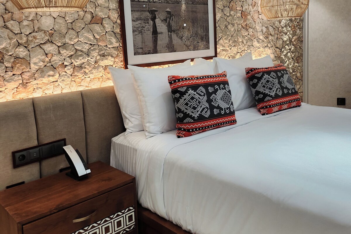 Quarto com parede de pedra, fotografia emoldurada, mesa de cabeceira com telefone e almofadas estampadas.