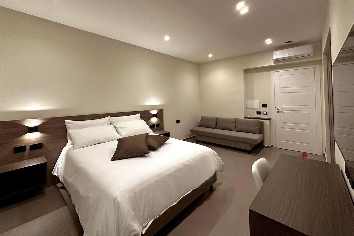 A modern hotel room with a large bed, bedside tables, a sofa, and a desk.