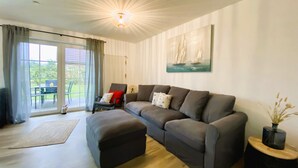A modern living room with a grey sofa, a matching ottoman, a dining table with chairs, and a large window with curtains.