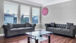En stue med to grå sofaer, et glasbord og store vinduer med persienner.