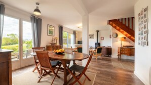 A dining area with a wooden table and chairs, a view through large windows, and a staircase leading to an upper level. - Holiday Home 'Maison Aux Volets Bleus - Sea View' with Private Terrace and Garden (Arzon)