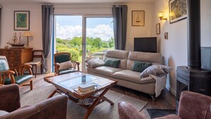 A living room with a sofa, chairs, a coffee table, a fireplace, and a large window with a view of greenery. - Holiday Home 'Maison Aux Volets Bleus - Sea View' with Private Terrace and Garden (Arzon)