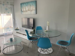 Un comedor moderno con una mesa de vidrio, sillas azules, un televisor y un cuadro decorativo en la pared.