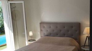 Una camera da letto con un letto grande, una testiera trapuntata, una lampada a sospensione, un comodino con una lampada e vista su una piscina attraverso una porta aperta.
