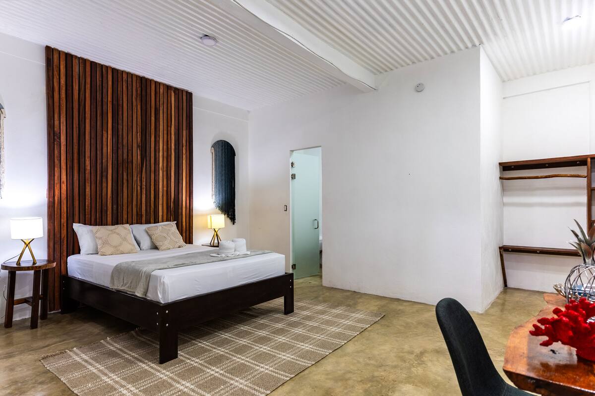 A modern bedroom with a wooden headboard, a bed with white linens, a bedside table with a lamp, and a small rug.