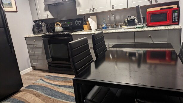 A modern kitchen with a black stove, a red microwave, and a stainless steel sink. There are two black chairs and a black table.