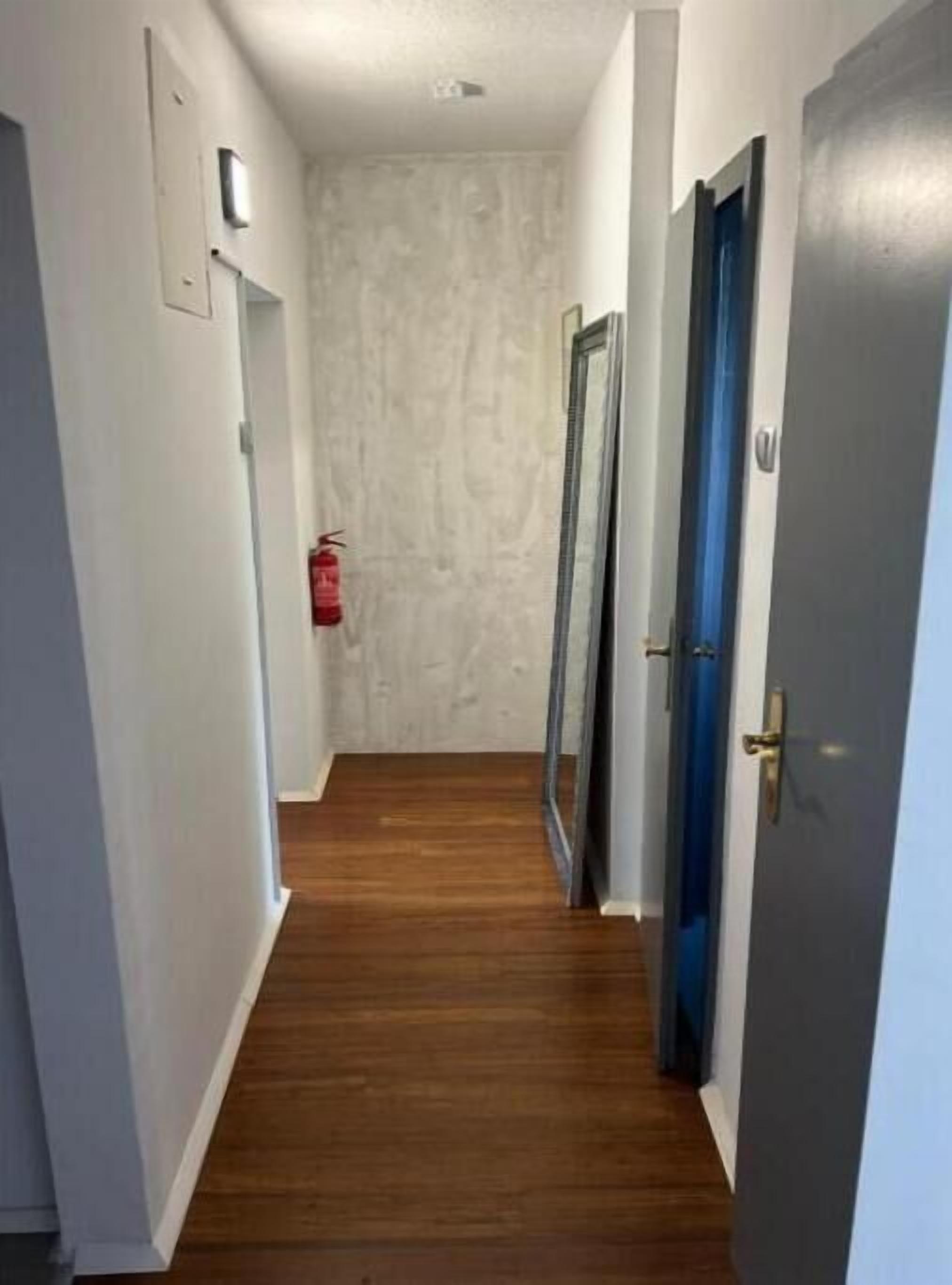 Un pasillo estrecho con suelo de madera, paredes blancas y una puerta azul a la derecha.