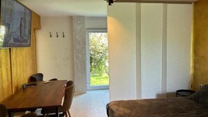 A modern bedroom with a large bed, a wooden dining table, and a view of a garden through a sliding door.