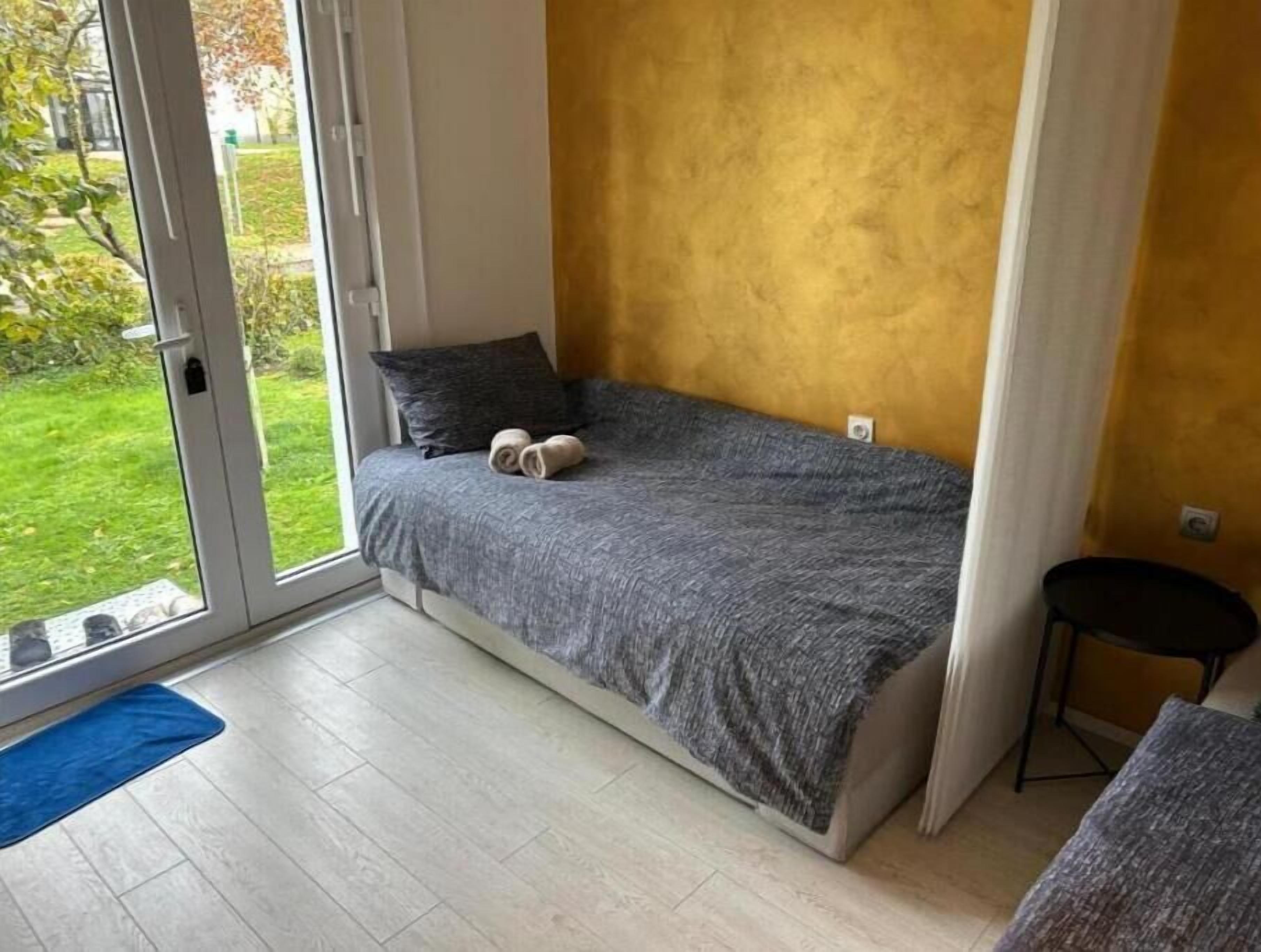 Ein kleines Zimmer mit einem Einzelbett, einer grauen Bettdecke, einem Paar zusammengerollter Handtücher, einem kleinen Hocker und Blick auf einen grünen Garten durch eine Schiebetür.