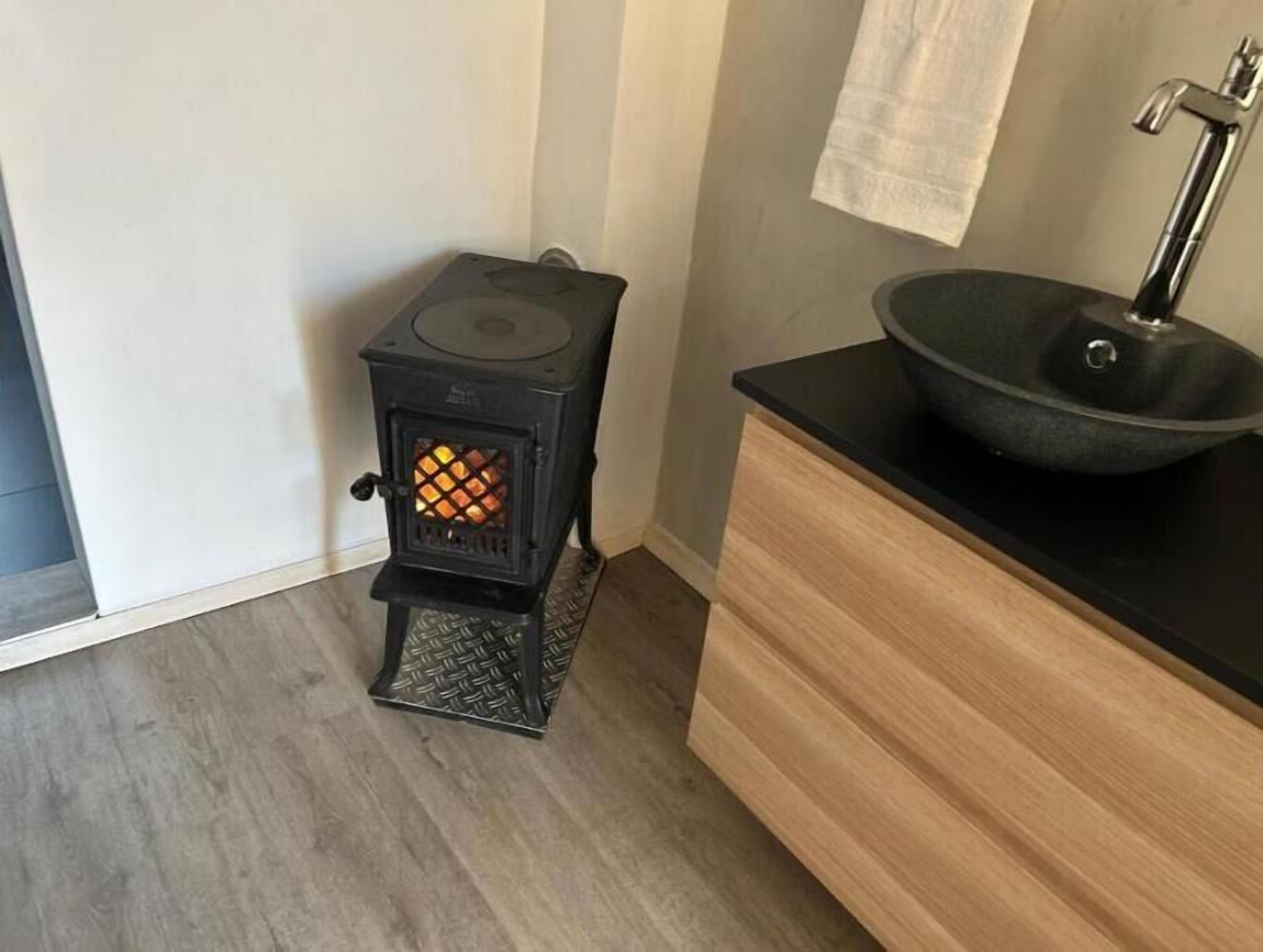Pieni, suljettu tila, jossa on pienikokoinen puukamiina, jonka liekit ovat näkyvissä, musta pesuallas ja puinen kaappi.