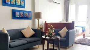 A modern hotel room with a blue sofa, a smaller armchair, a round table with a vase of flowers, a bed with a wooden headboard, and artwork on the wall. - Merci Buccoo (Scarborough)