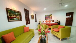 A hotel room with a green sofa, a wooden coffee table with a vase, and a bed with orange pillows. - Merci Buccoo (Scarborough)