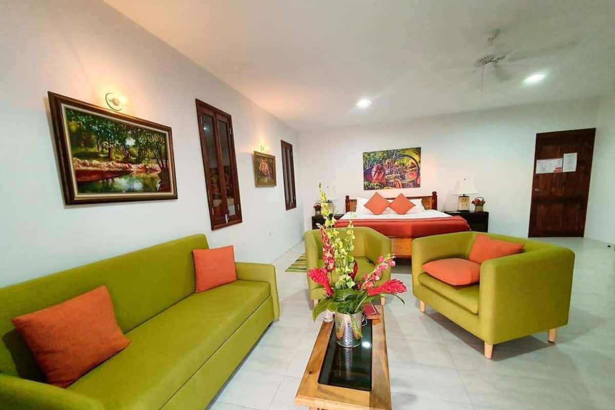 A hotel room with a green sofa, a wooden coffee table with a vase, and a bed with orange pillows.