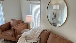 A brown leather sofa with a throw blanket and a pillow with the text "be our guest" in a room with a large mirror, a floor lamp, and a window with blinds.