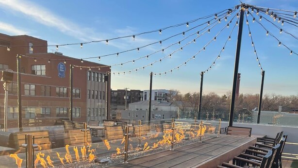 A long wooden bench with metal frames, a fire feature with orange flames, and string lights overhead.