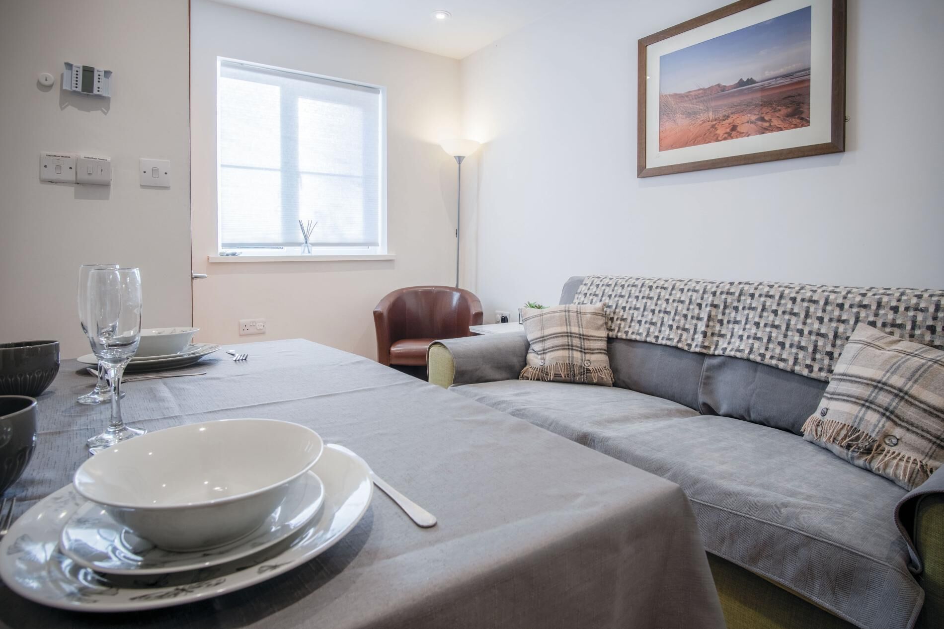 Un salon moderne avec un canapé gris, une table à manger dressée avec des assiettes et des verres, une fenêtre avec des stores et un tableau encadré au mur.